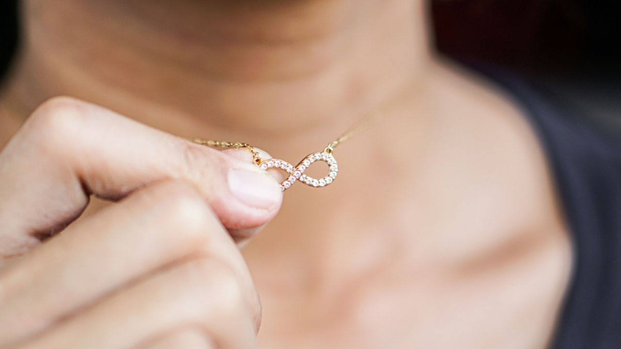 Close-up of a person's hand holding a delicate yellow gold infinity pendant necklace accented with pavé diamonds.
